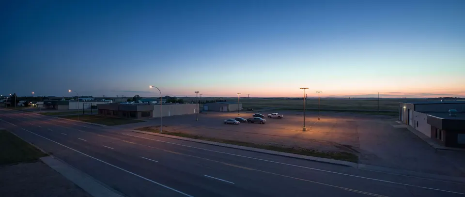 A wide commercial avenue at blue hour on the edge of a prairie city, a parking lot under tall lamps, flat land and a pale band of light on the horizon.